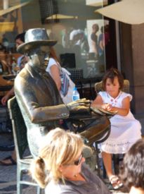 En jente setter seg på den ledige bronsestolen og tilbyr ham noe å drikke. Ungdommer klapper ham på hatten idet de passerer. Det ser ut som bronsestatuen tiltrekker seg trøstens gest. På kvelden er han omringet av originaler, musikere og turister. 
foto©Tove Andersson 